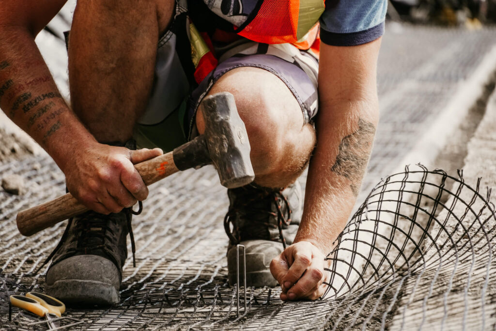 Close-up of an Earth and Turf Landscaping Edmonton worker hammering pegs into wire mesh on a retaining wall project.