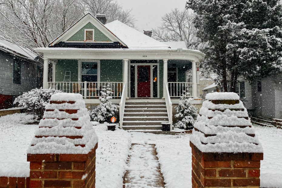 Snow is falling onto a snow covered lawn and home.