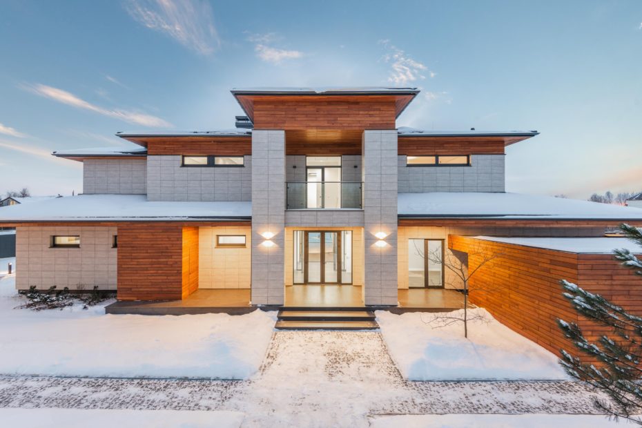 A large, modern style home is snow covered and brightly lit.